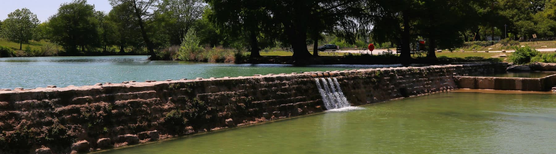 A low stone dam on the Blanco River with water spilling over it, under cypress and live oaks